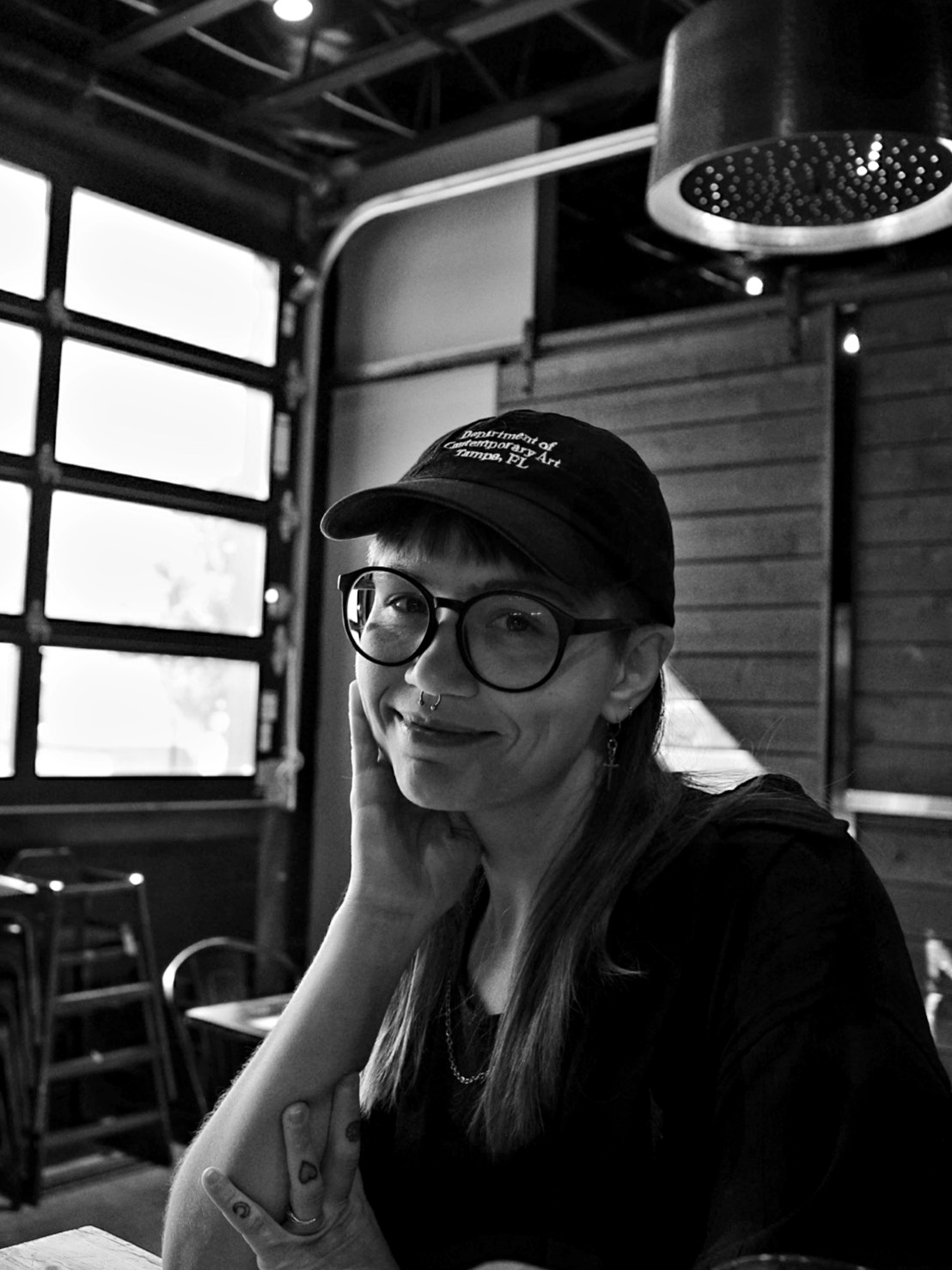 a photo of mia at a table. they are wearing a Department of Contemporary Art hat and their hand is resting on their cheek.